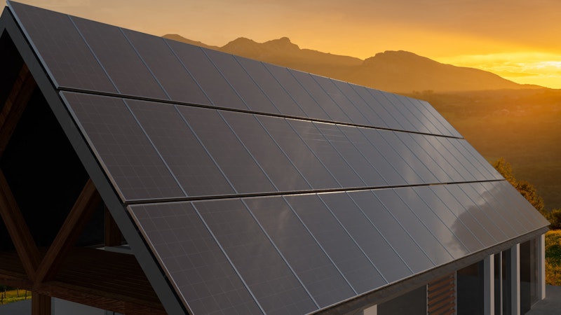 Black solar panels set on a steap angled roof backlit by the sunest over the mountains