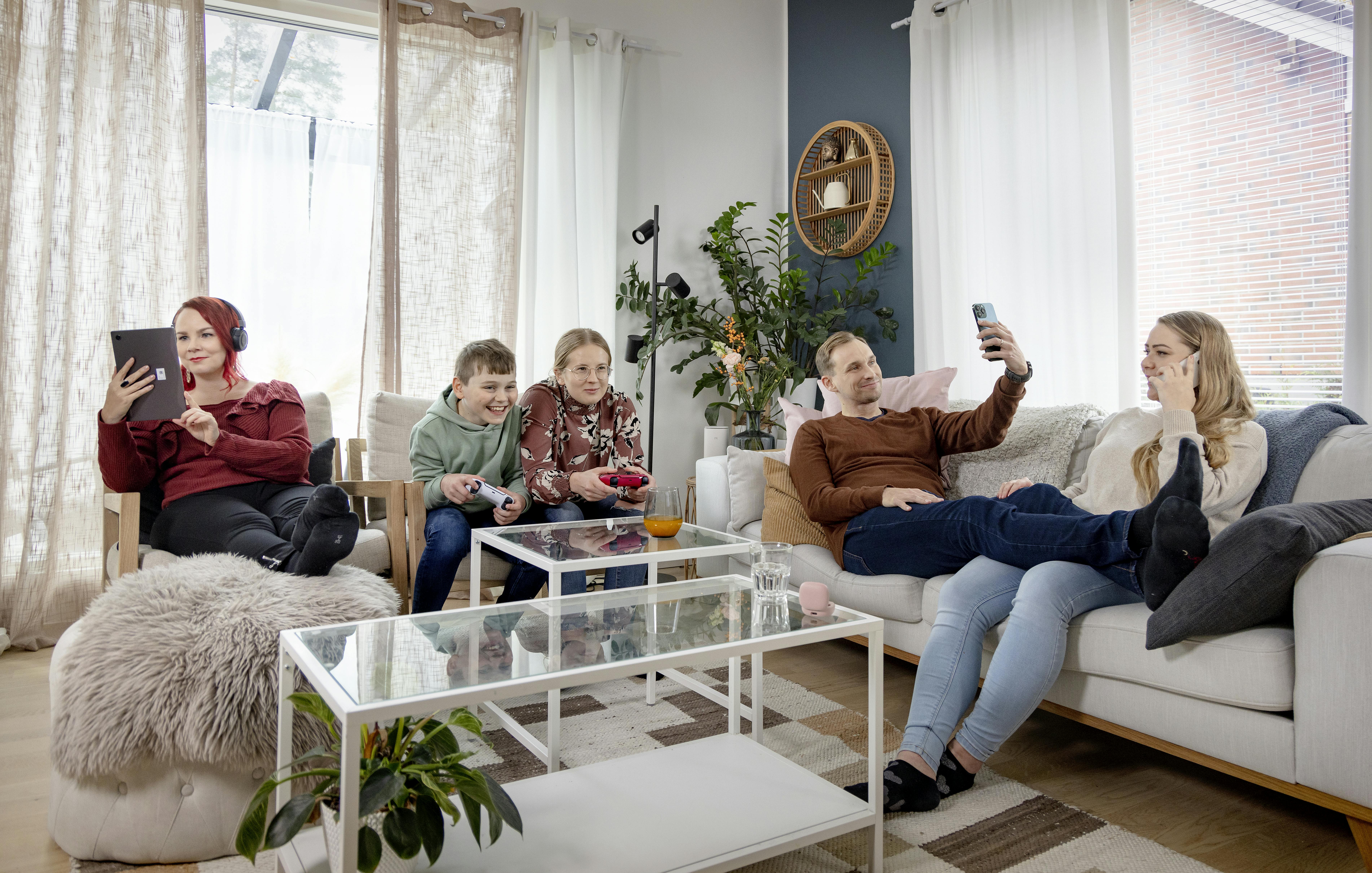 3 lasta ja 2 aikuista käyttävät eri elektroniikkalaitteita olohuoneessa.