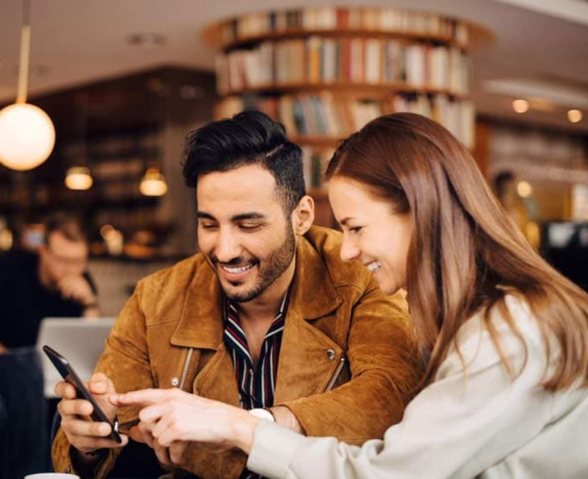 A man and woman looking at a mobile phone in a lounge scrolling through the Amex MENA website.