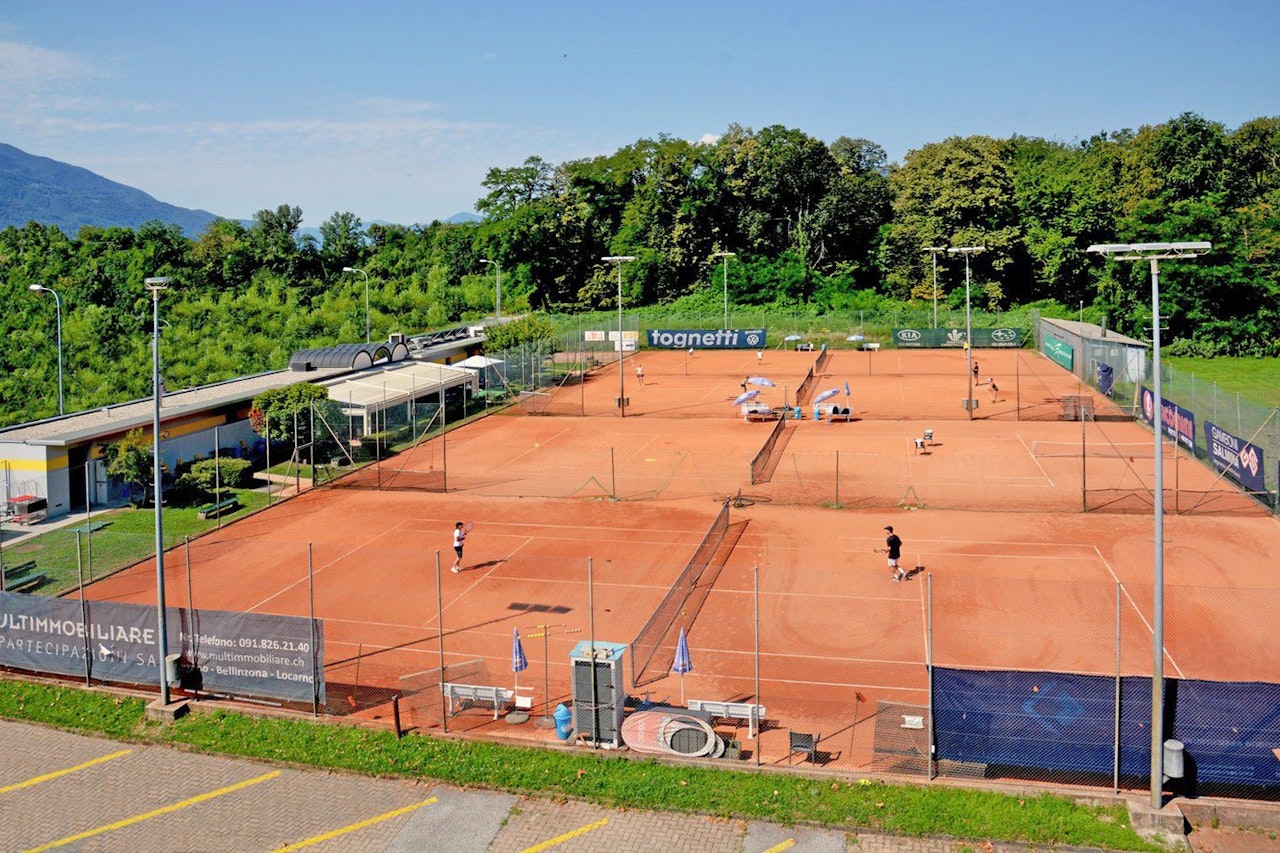 Ein großer Tennisplatz aus Sand mit mehreren spielenden Personen unter klarem Himmel, umgeben von üppig grünen Bäumen und Bergen in der Ferne, ideal für Tennis im Freien.