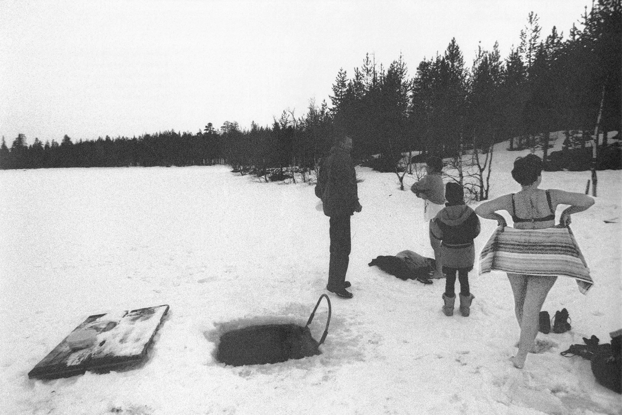 Svartvitt foto av fyra personer vid ett hål i isen för isbad, två av dem på väg att klä av sig.