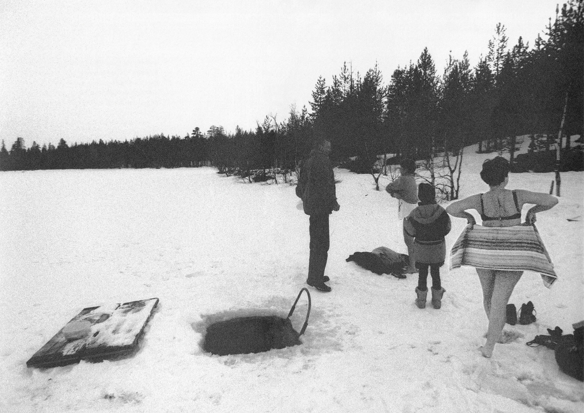 Svartvitt foto av fyra personer vid ett hål i isen för isbad, två av dem på väg att klä av sig.