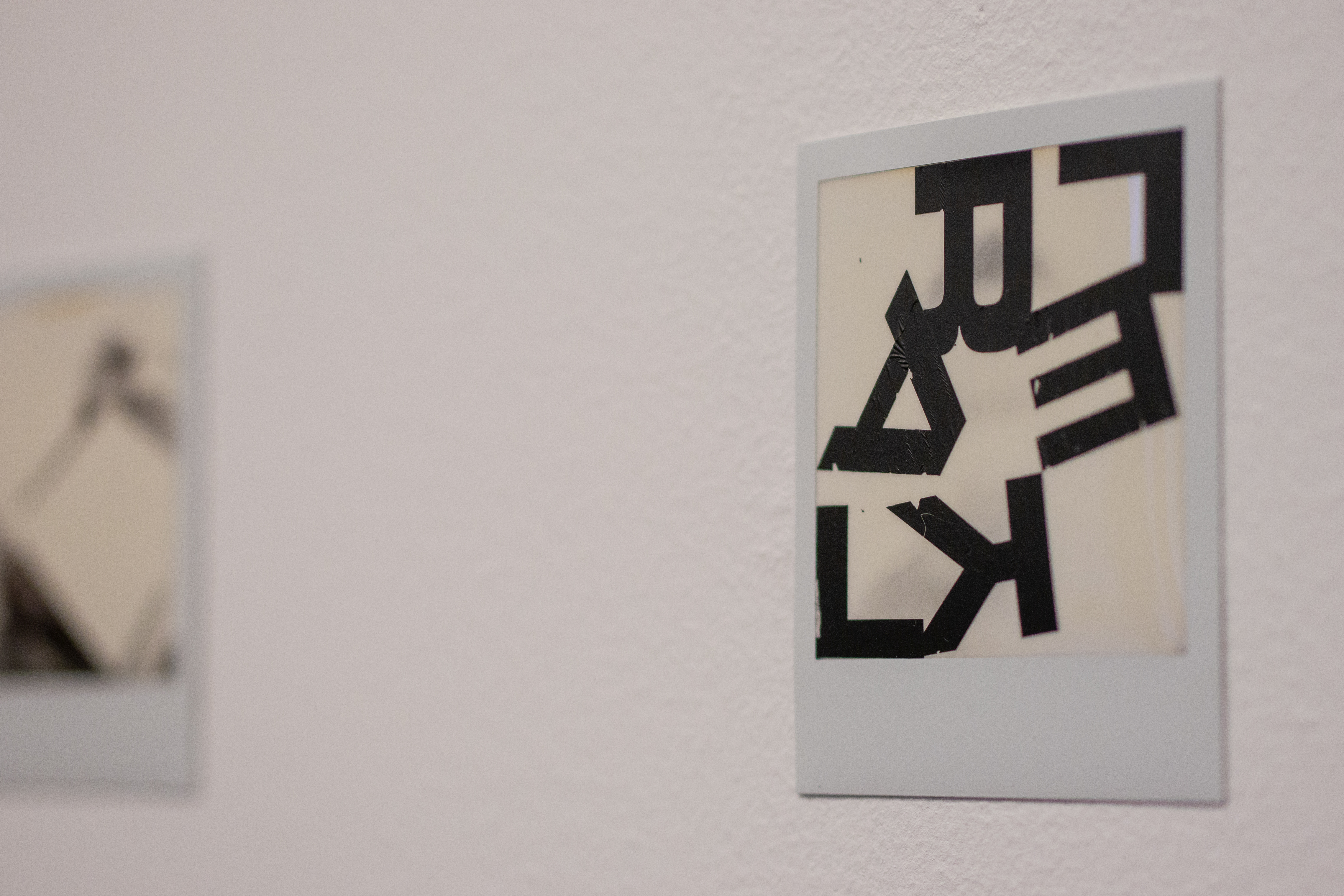 Närbild på ett polaroidfotografi fäst på en vit vägg, med ett suddigt ytterligare fotografi i bakgrunden. Det går inte att urskilja vad fotografierna föreställer, men det i förgrunden är bearbetat, med bokstäver som t.ex. L, E, A, K, F klistrade ovanpå bilden. Bokstäverna bildar inga urskiljbara ord.