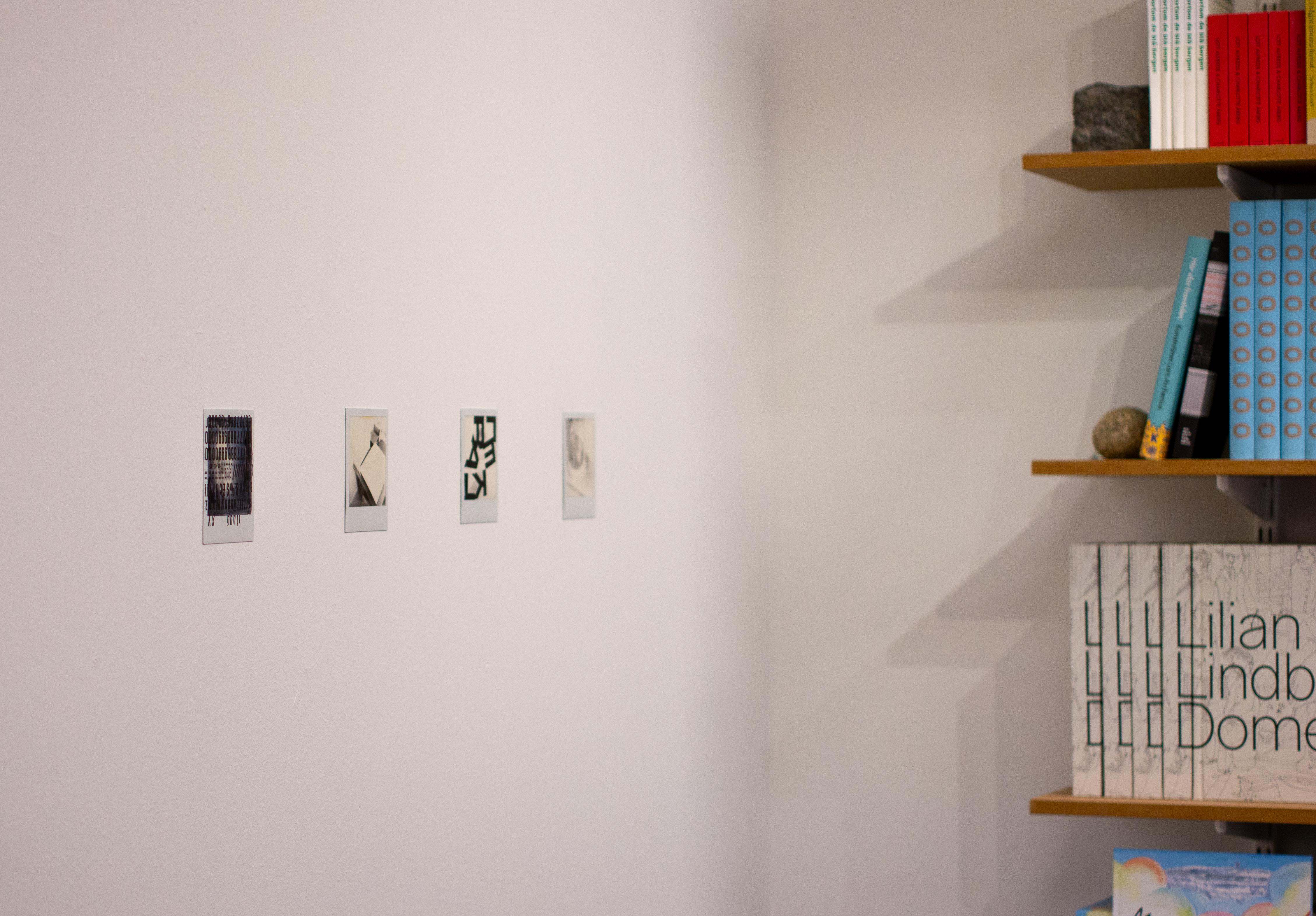 Fyra polaroidfoton sitter på rad på en vit vägg till vänster, till höger syns en bokhylla. Det går  inte att urskilja vad fotografierna förställer, men enstaka påklistrade bokstäver kan synas, till exempel F, L och K. De bildar inga urskiljbara ord.