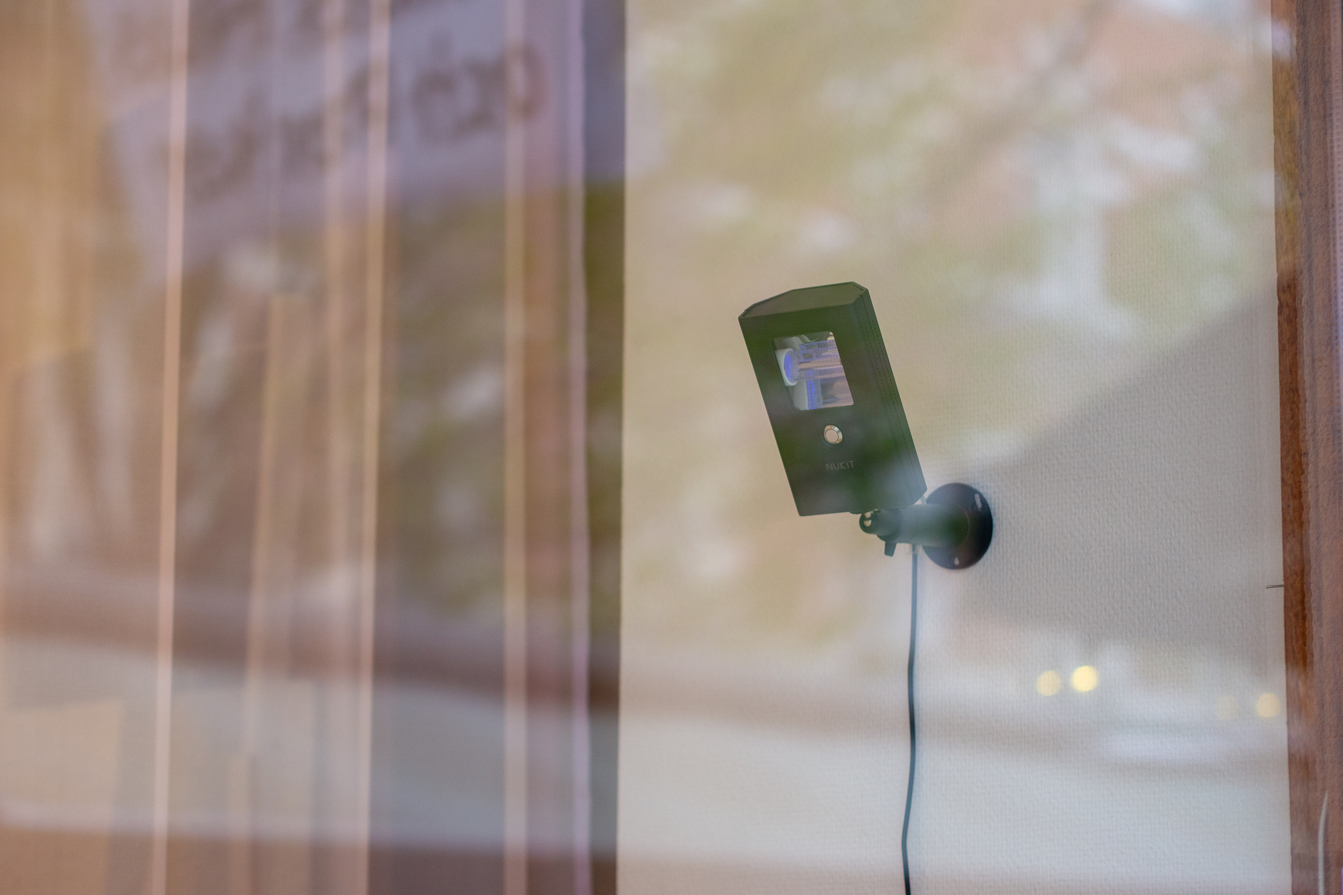 En svart rektangulär lampa med blått ljus sitter fäst på en vit vägg, fotat genom ett fönster. I glasets reflektion syns gatan utanför.