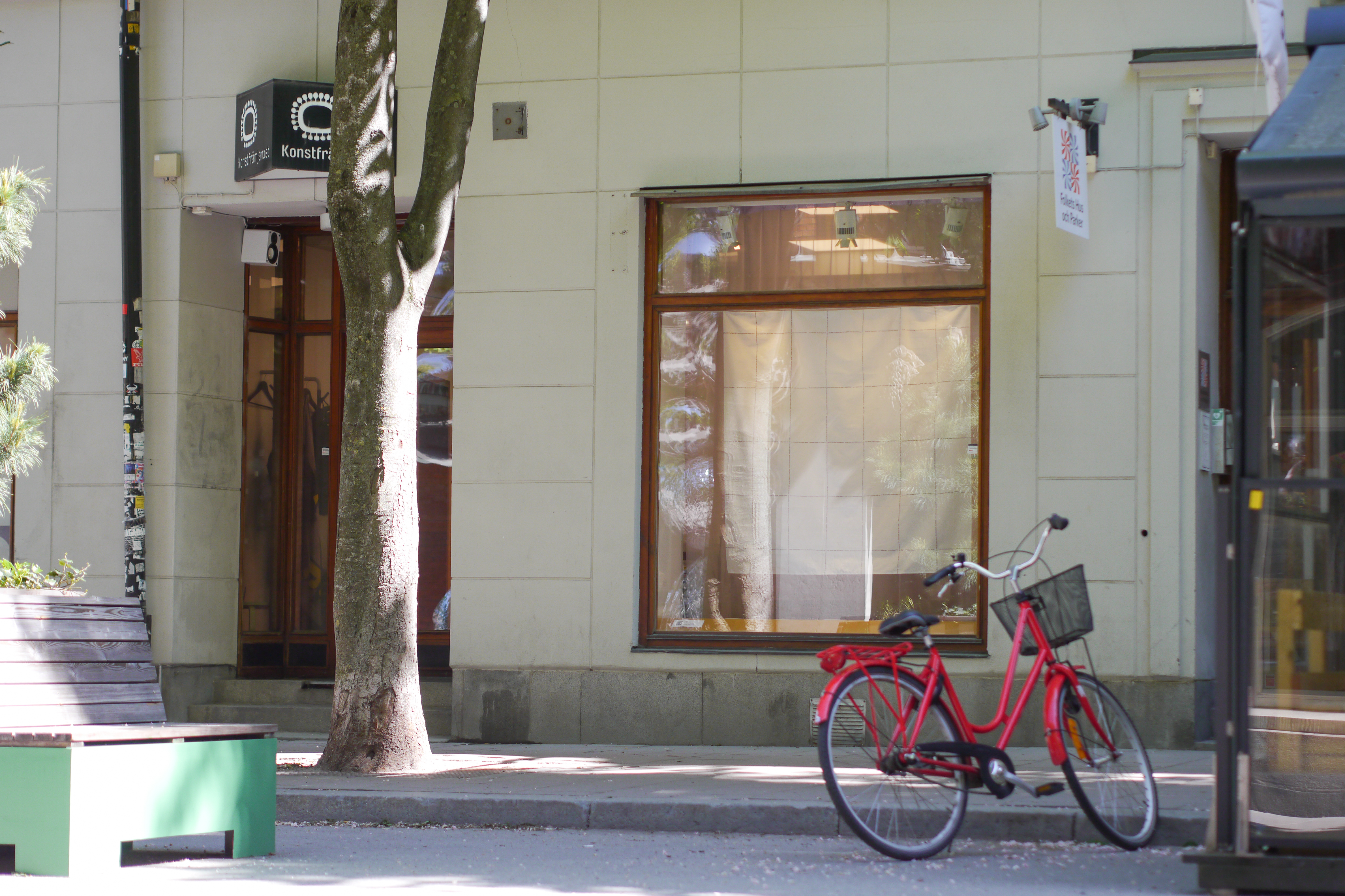 Foto av Konstfrämjandets skyltfönster och entré på markplan på Swedenborgsgatan i Stockholm, en solig dag. I förgrunden syns en  bänk i trä, ett träd och en röd cykel.  I skyltfönstret hänger en 150x150 cm stor vit textil med tunt rutmösnter i svart.