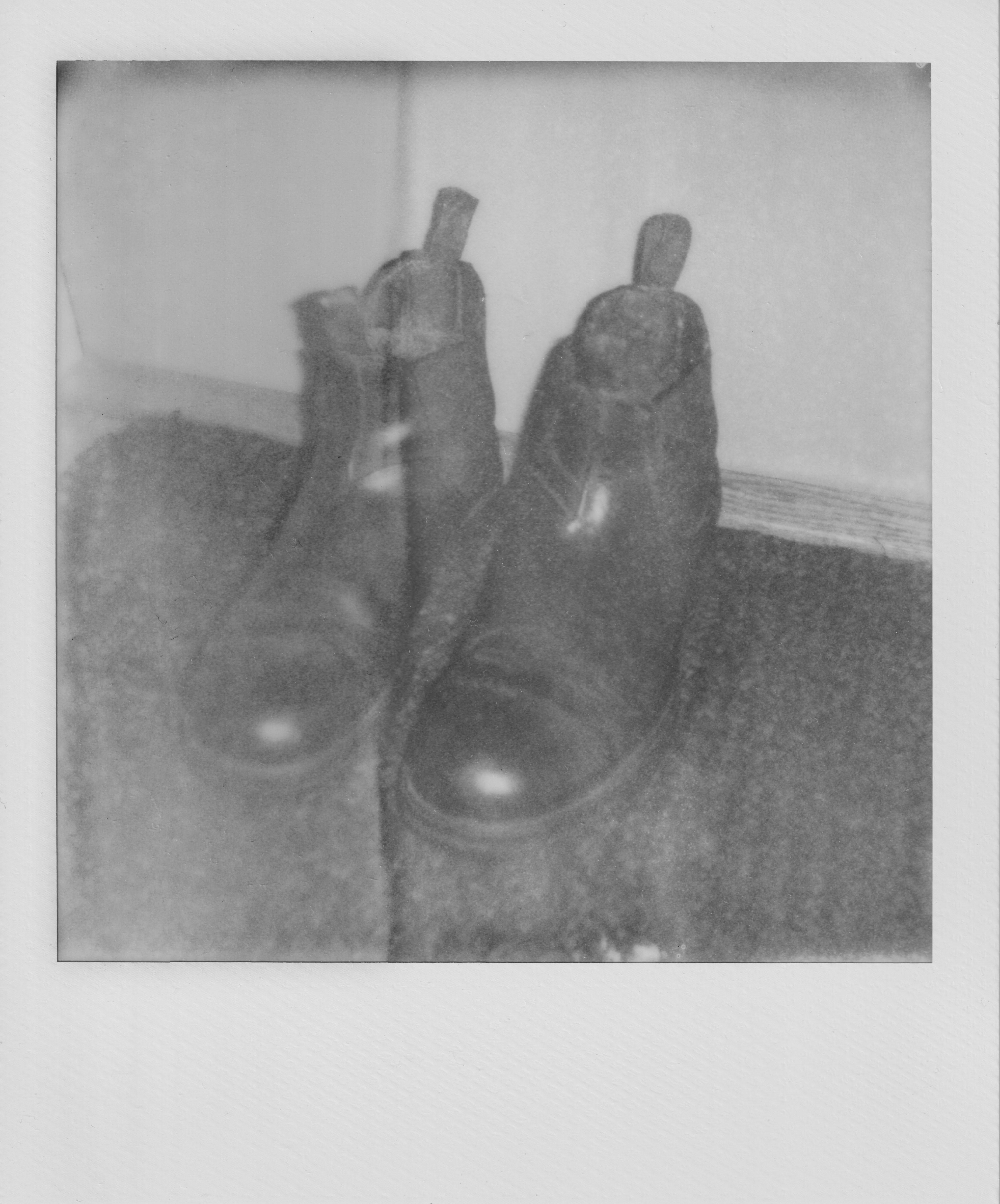 Ett svartvitt polaroidfoto av ett par svarta vinterstövlar placerade på en dörrmatta intill en ytterdörr. Fotot är något suddigt och har exponerats felaktigt så att det går en vertikal spricka igenom hela bilden, vilket ger bilden ett åldrat intryck. Fotot är ett verk av konstnären Diktgymnasiet och har titeln Monument 20250826.