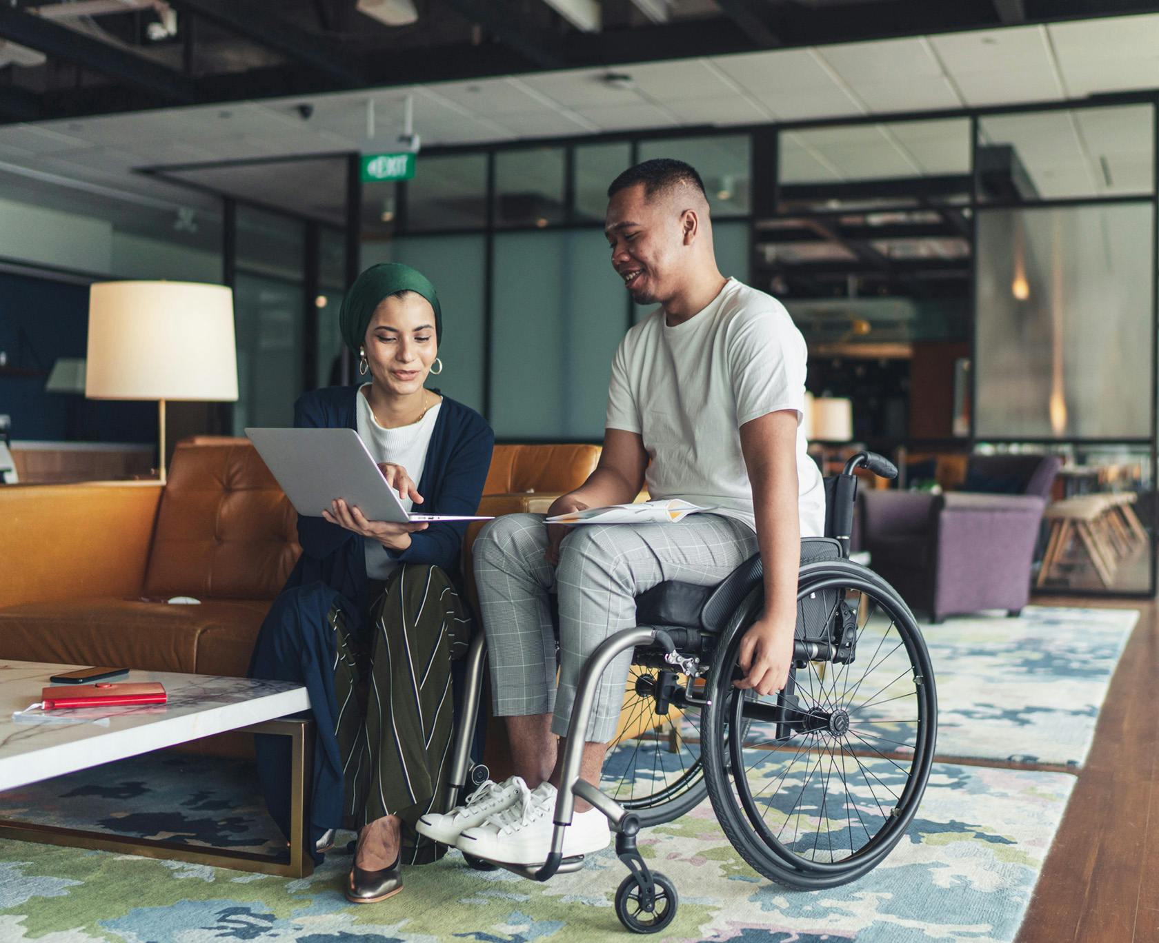 Man in a wheel chair looking at a computer with a woman sitting on a couch