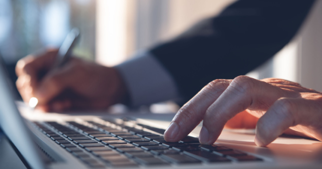 A man using a laptop and writing down notes as he calculates the cost of recruitment