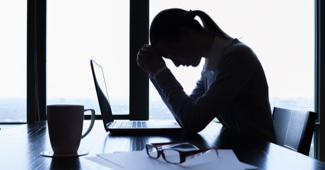 A woman in shadow sitting at a desk with her head in her hands