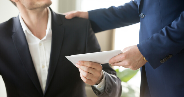 A male manager handing an male employee an envelope containgin a redundancy notification letter.