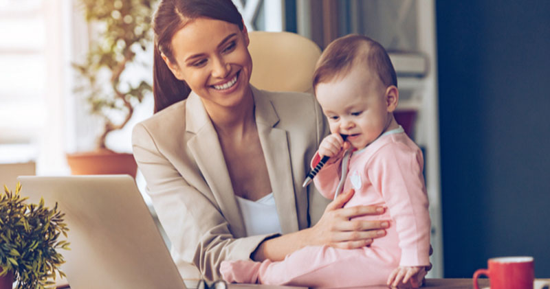 young mother plays with baby daughter while remote working at laptop