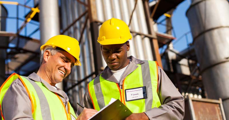 two male workers in high vis look at charts outside large factory