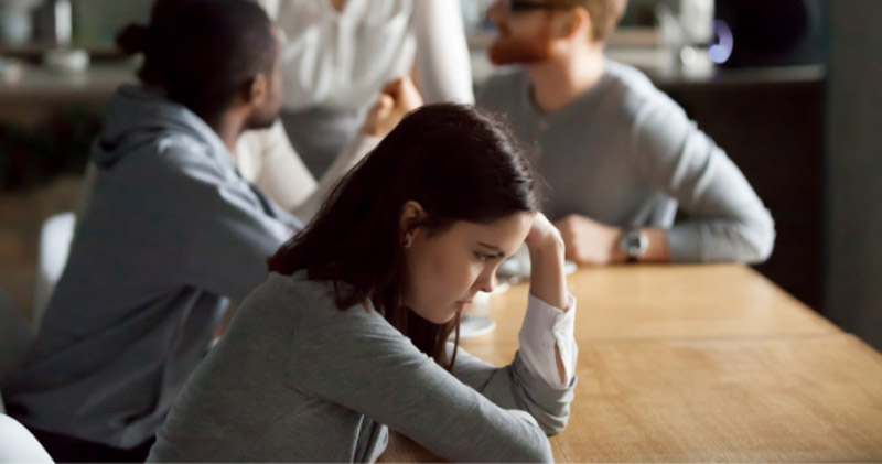 isolated female employee sits away from male employees in dark lunchroom