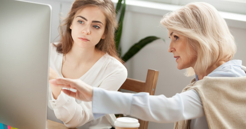 older female employee assists younger female employee with computer