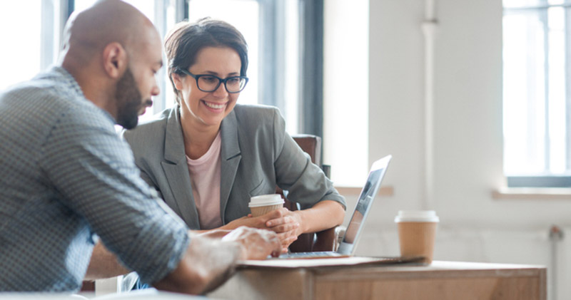 man and woman look at laptop together in sunlit office