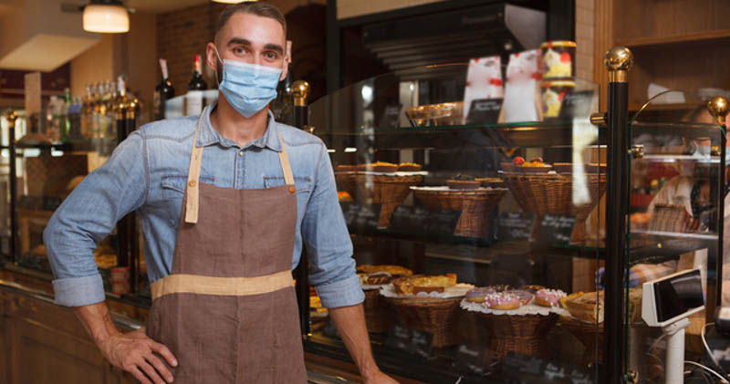 cafe owner smiles with face mask in front of cake window
