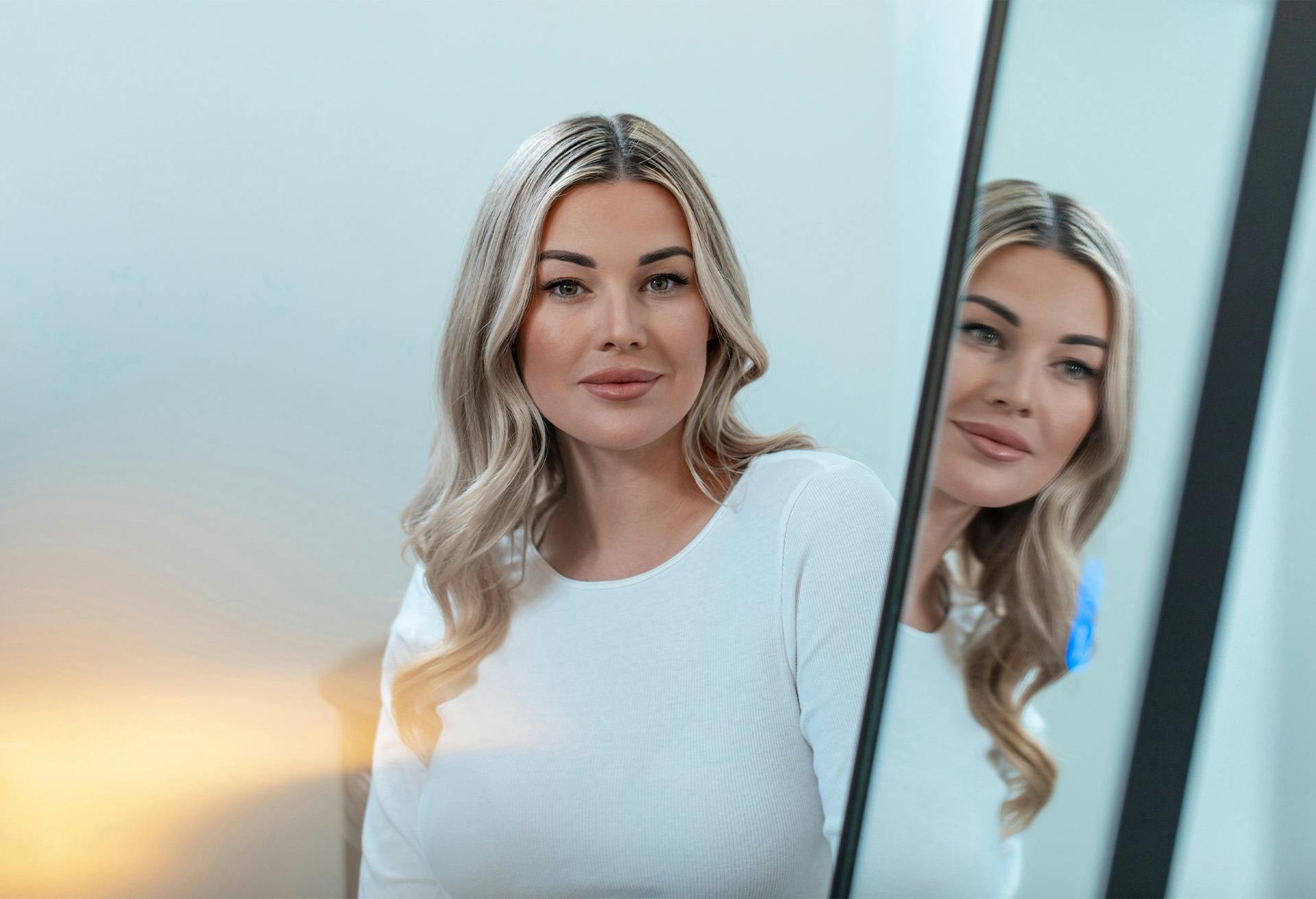 A blonde woman in white standing by a mirror