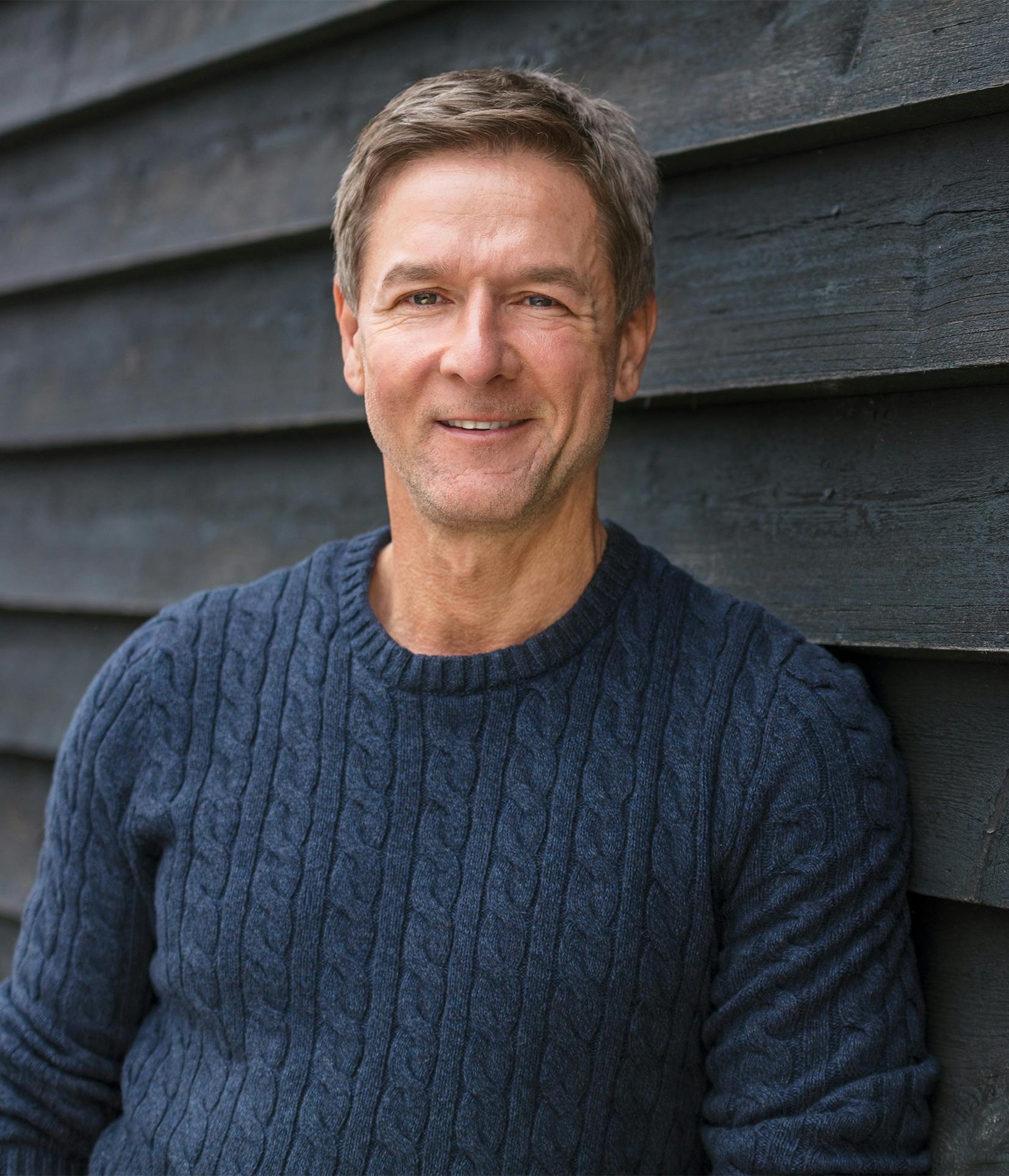 smiling man in blue sweater leaning against a black wall