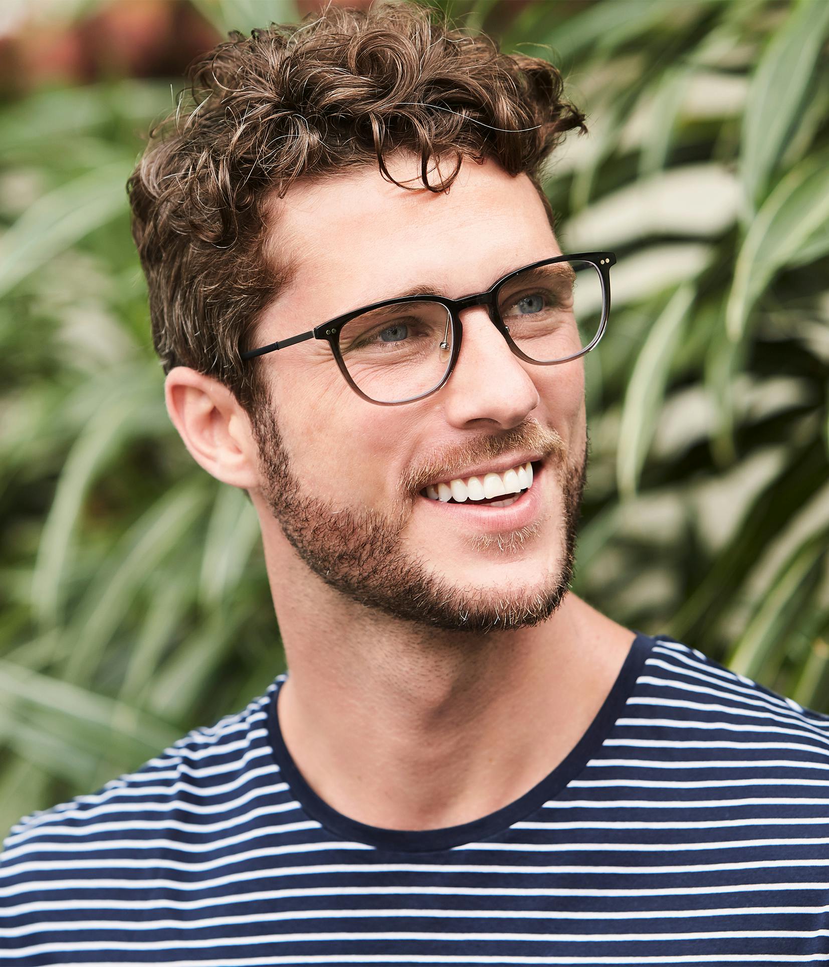 smiling man with glasses and a striped shirt in front of a bush