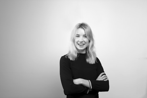young woman in her 20s or 30s with shoulder length blonde hair, standing with her arms crossed, looking to camera and smiling
