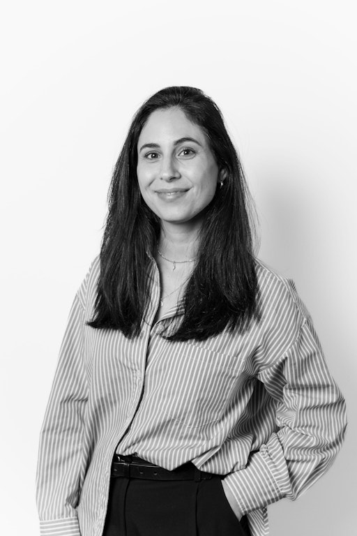A woman with long dark hair wearing a striped shirt and dark trousers stands against a plain background, smiling at the camera.