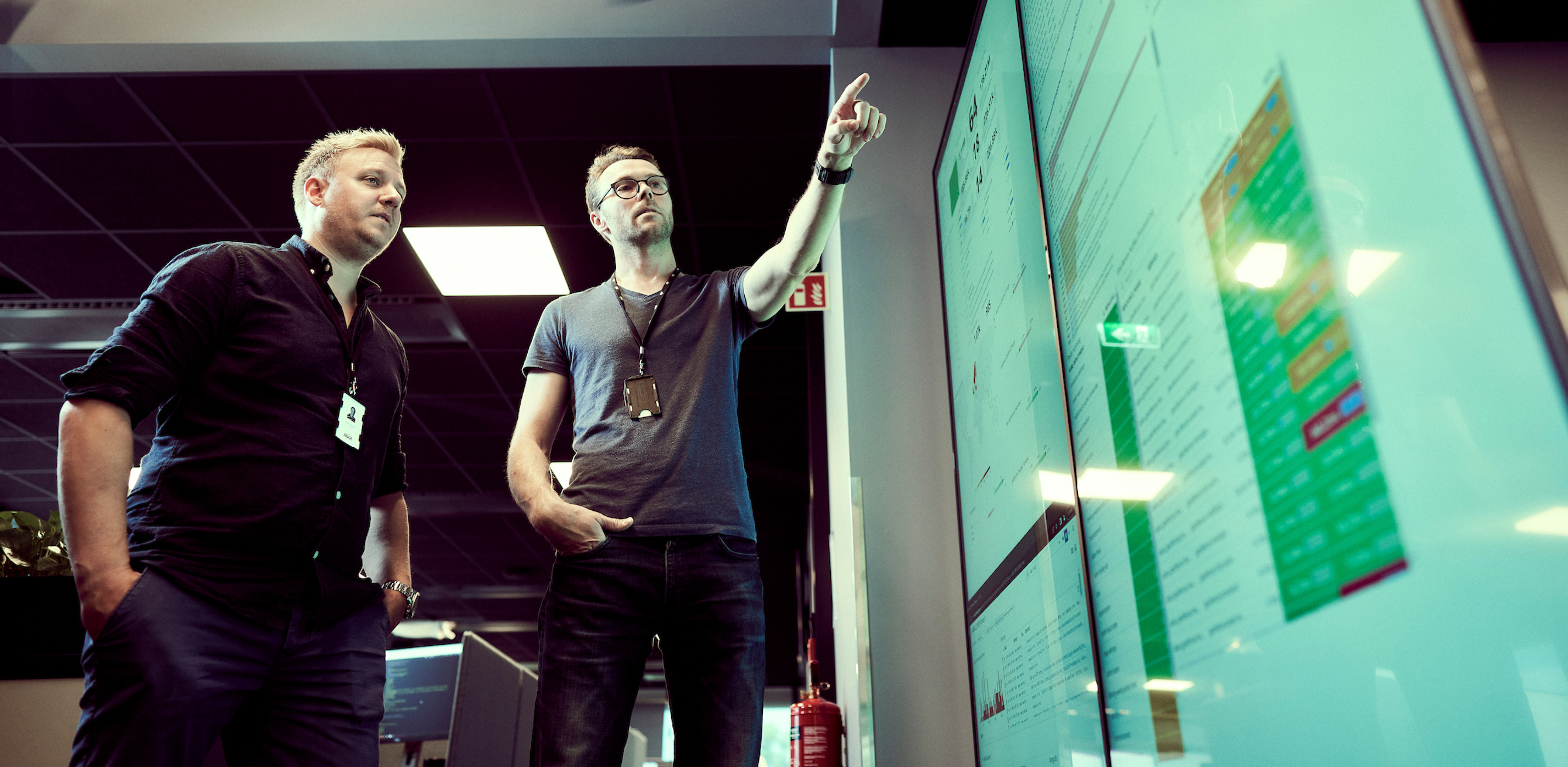 Two men in black tshirts pointing towards data displayed on large screen