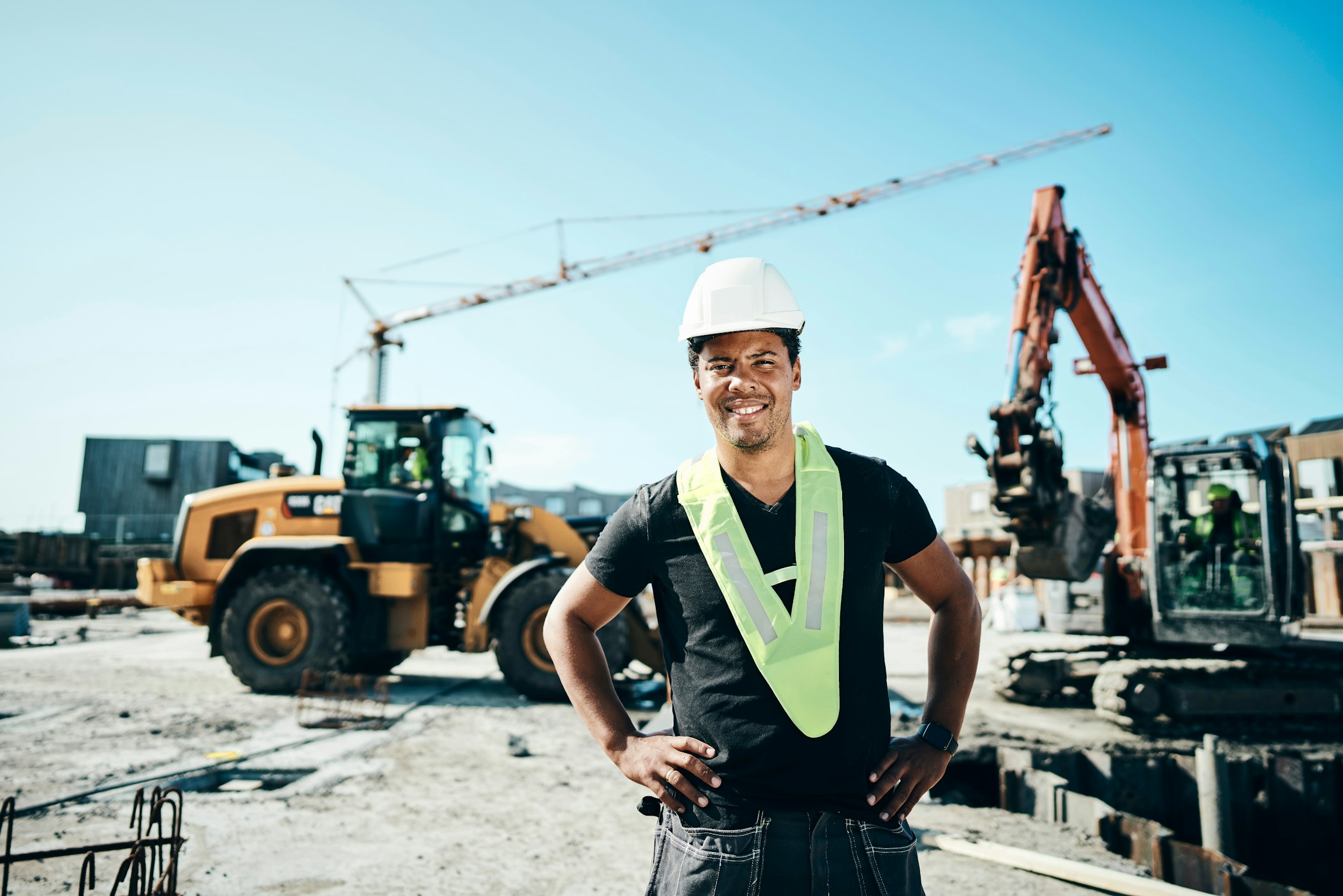 Een medewerker in fluohesje en veiligheidshelm poseert voor graafmachines en andere apparatuur.