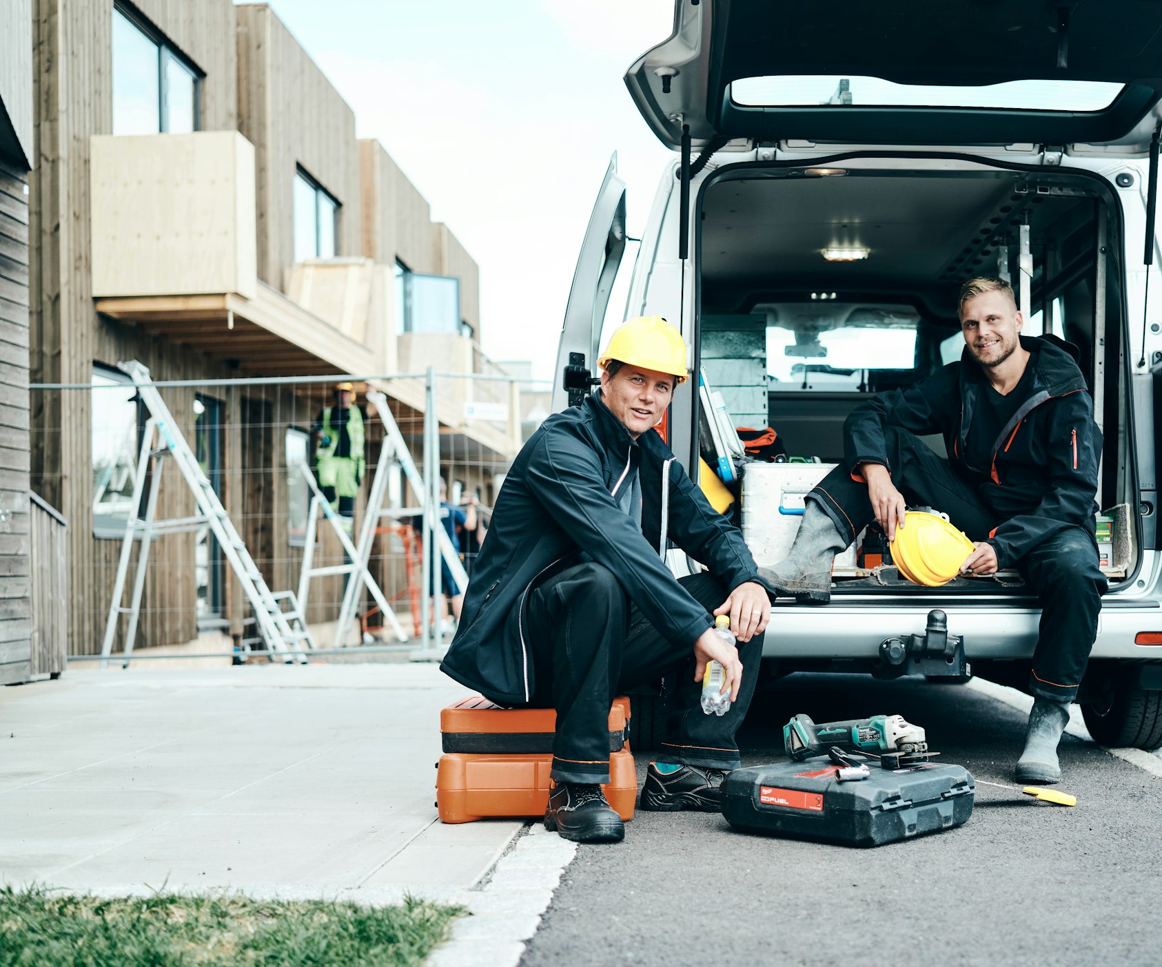 To bygningsarbejdere holder pause og sidder bag en varevogn med værktøj på gulvet.