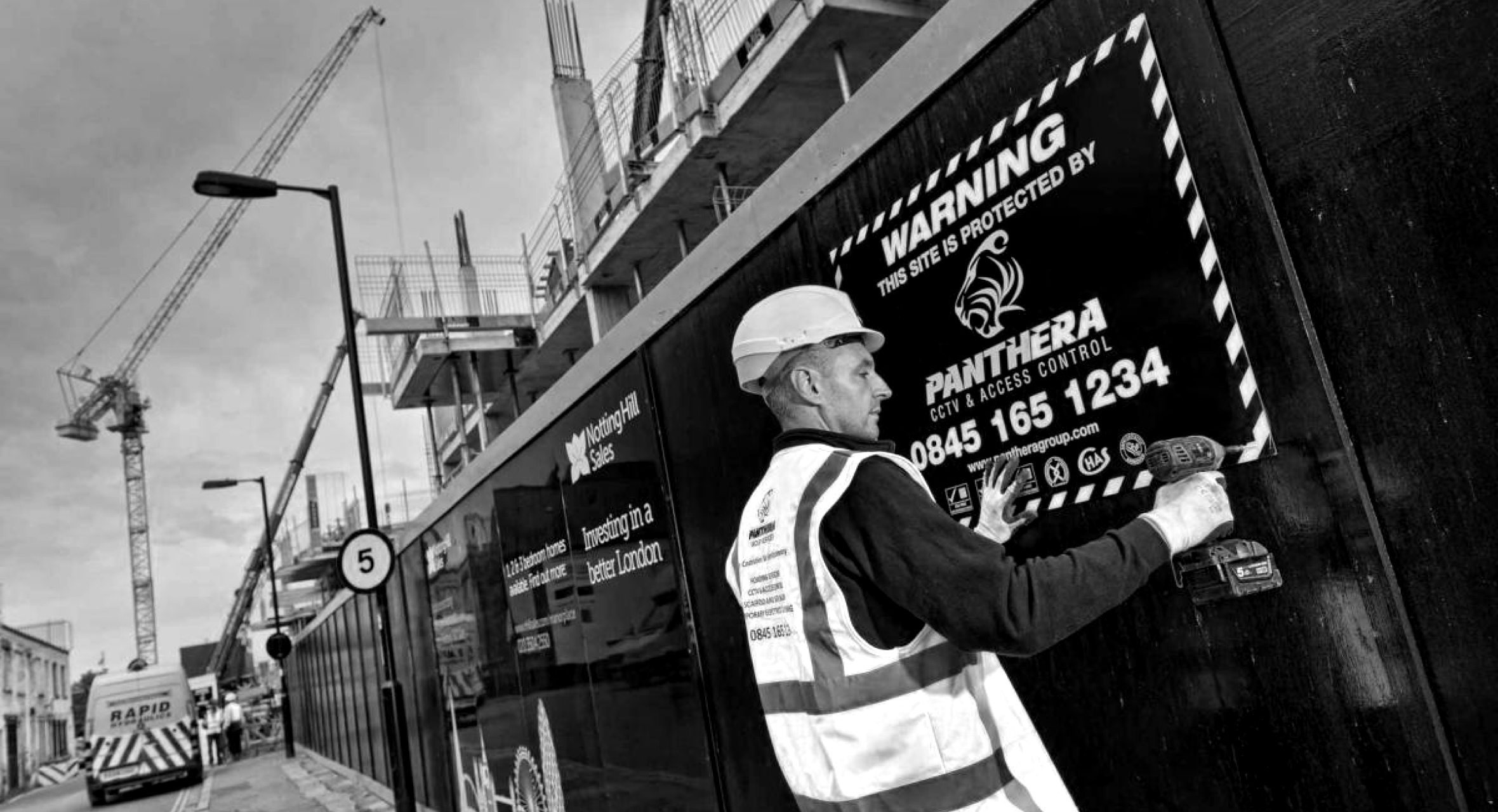 Black and white image with man in construction safety clothes putting up Panthera Group sign on construction site fence fence,