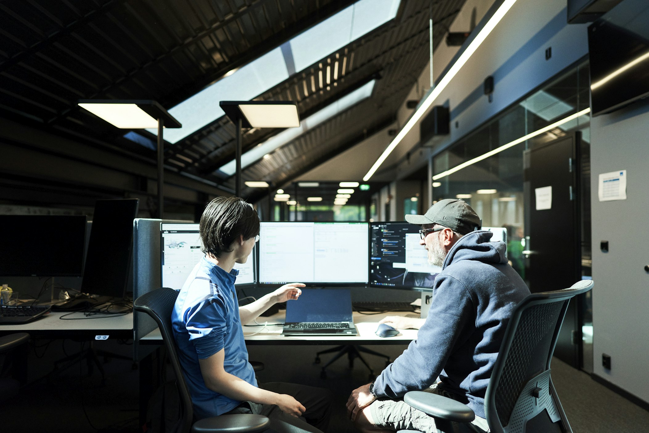 Two people sit at a desk in a modern office, looking at computer monitors with code and documents displayed. One person points at a screen while the other listens, suggesting collaboration or discussion.