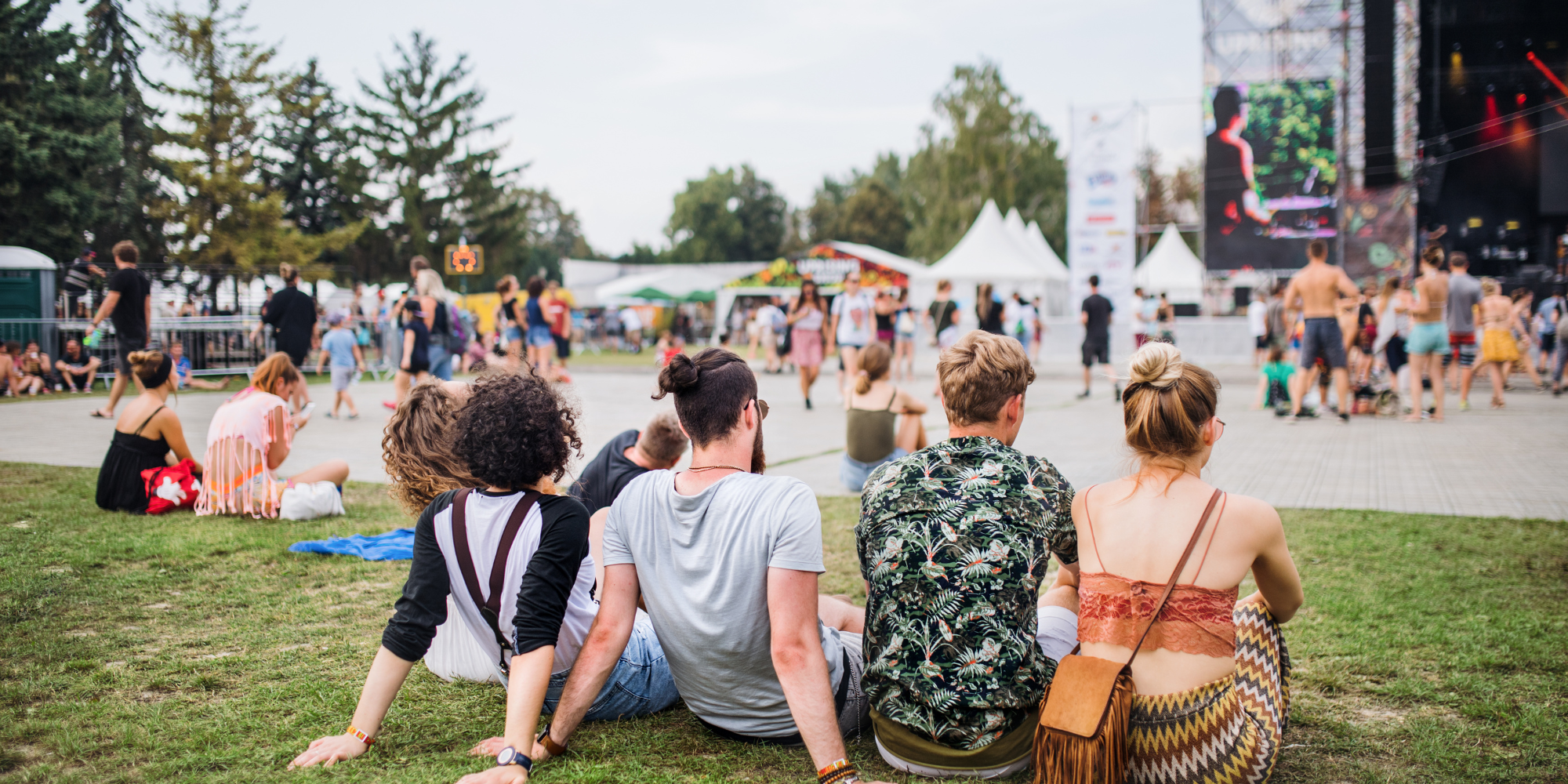 5 people sitting on the grass at a festival watching a band on the stage