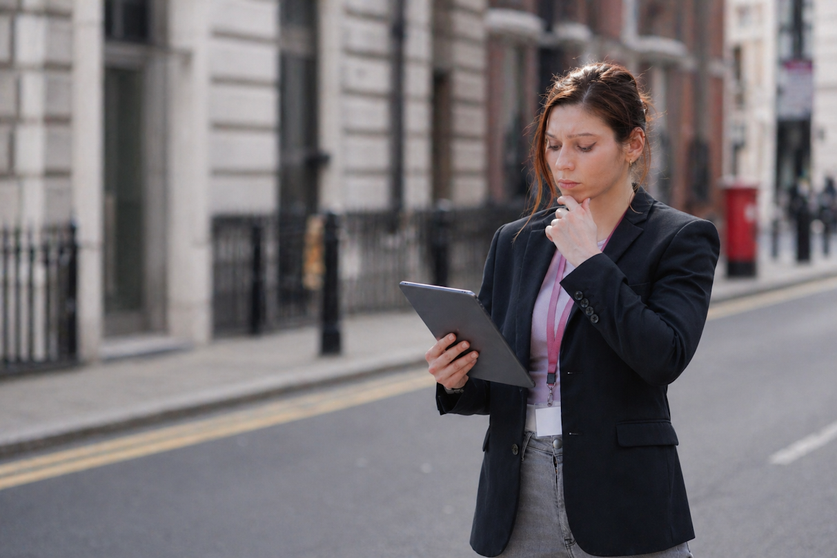 Young woman in dark blazer and lanyard staring thoughtfully at tablet