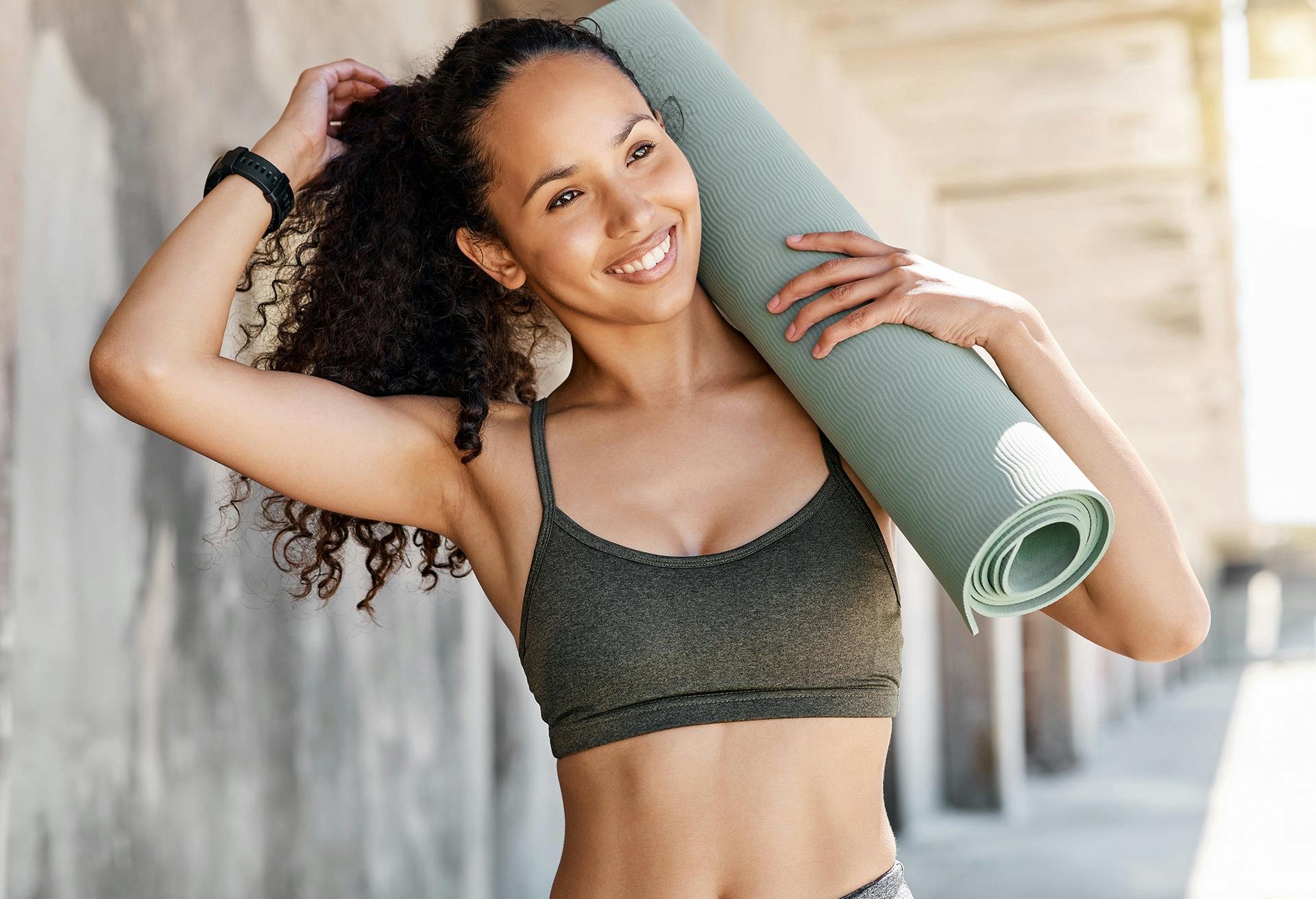 Woman outside smiling while wearing a workout top and carrying a yoga mat