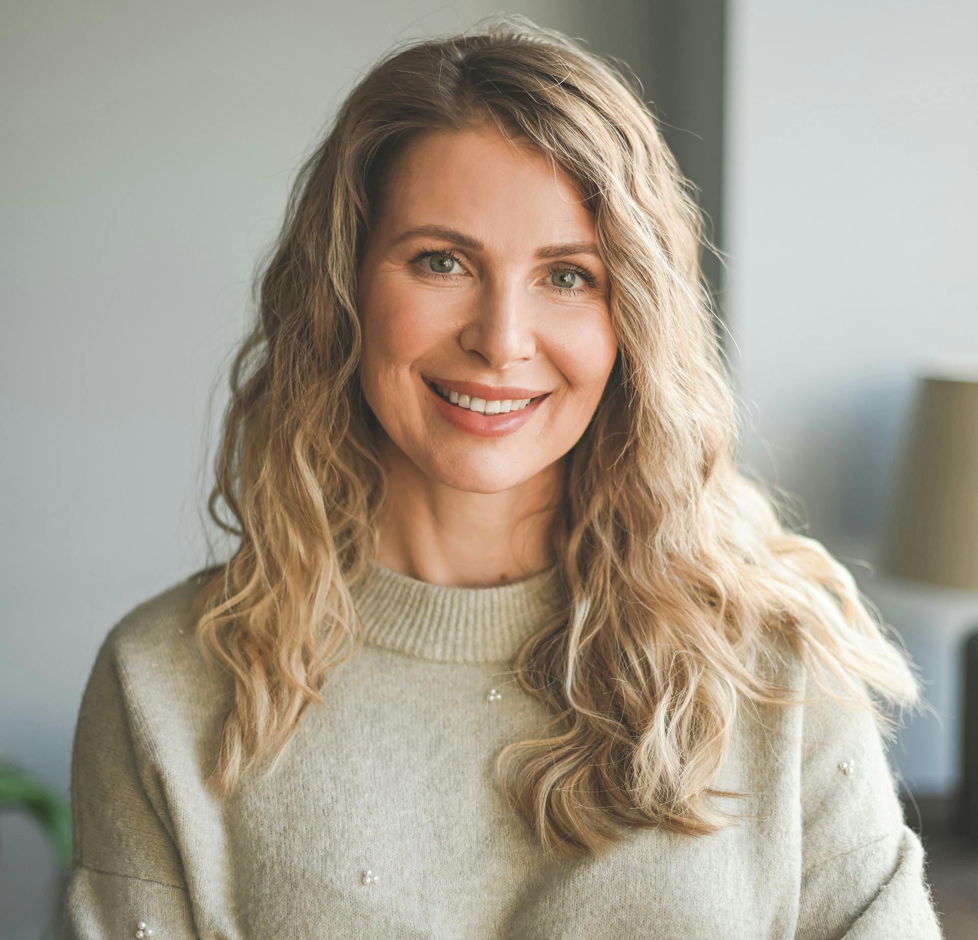Woman with long curly blonde hair smiling and wearing a cream colored sweater
