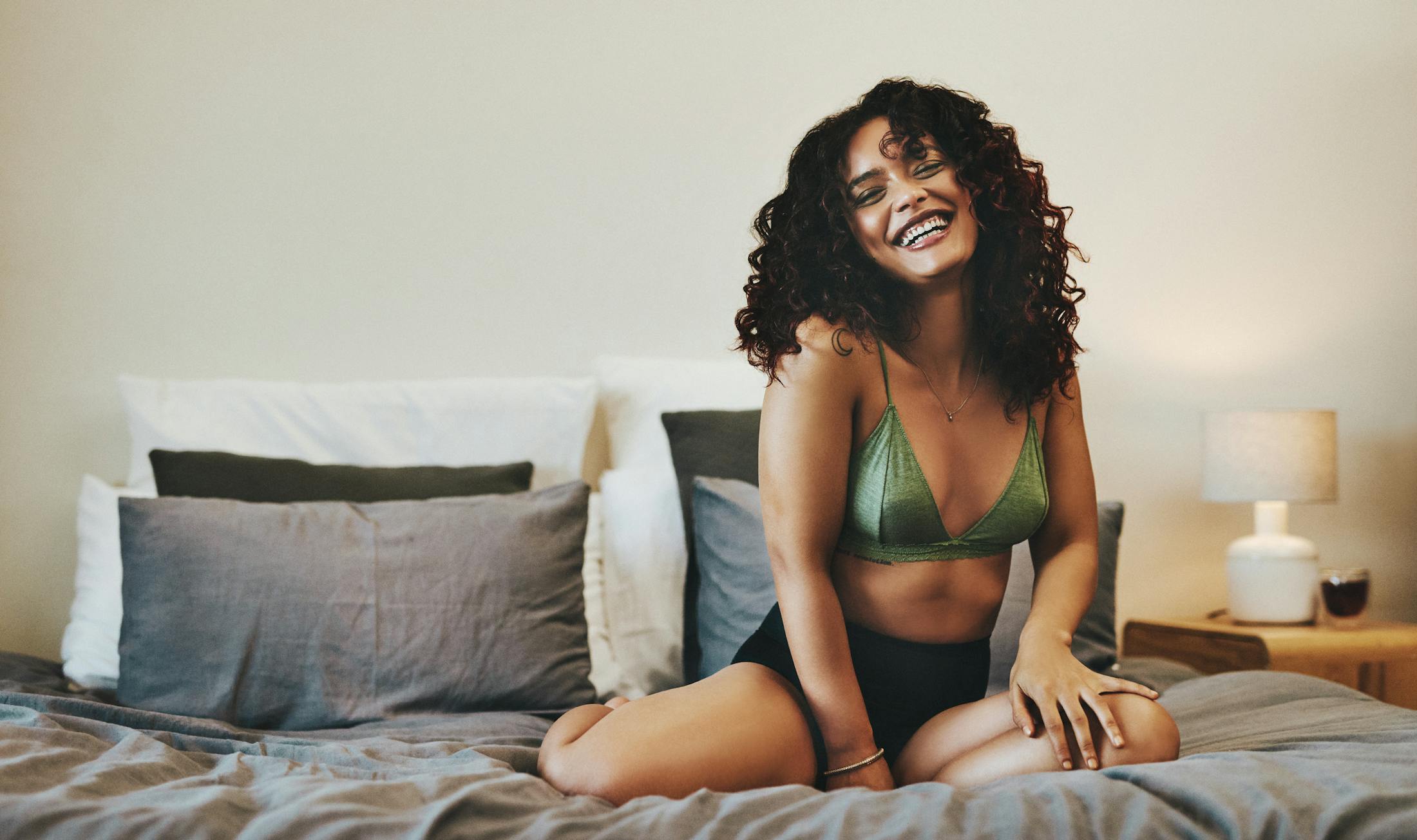 Woman with curly hair laughing while sitting in bed in her underwear