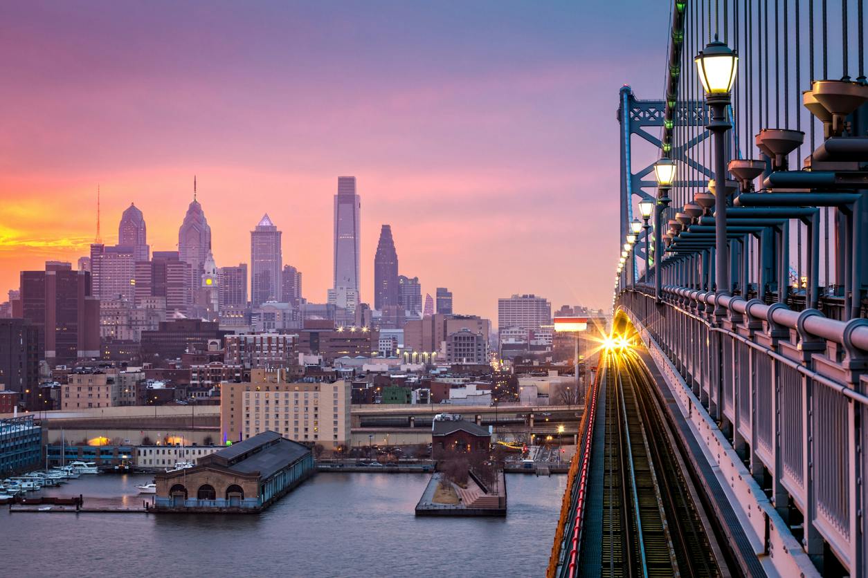 Philadelphia under a hazy purple sunset. An incoming train crosses Ben Franklin Bridge.