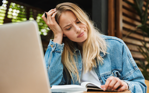 Woman sits in front of laptop while looking at a book.