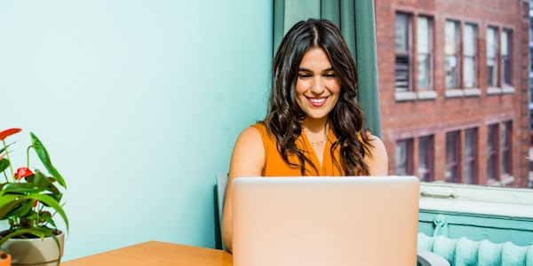 A woman sitting in a well lit room by the window, looking cheerfully at a laptop.