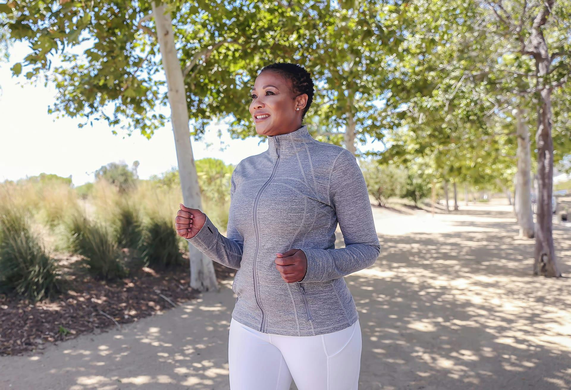 smiling woman in grey jacket and white pants jogging on a path