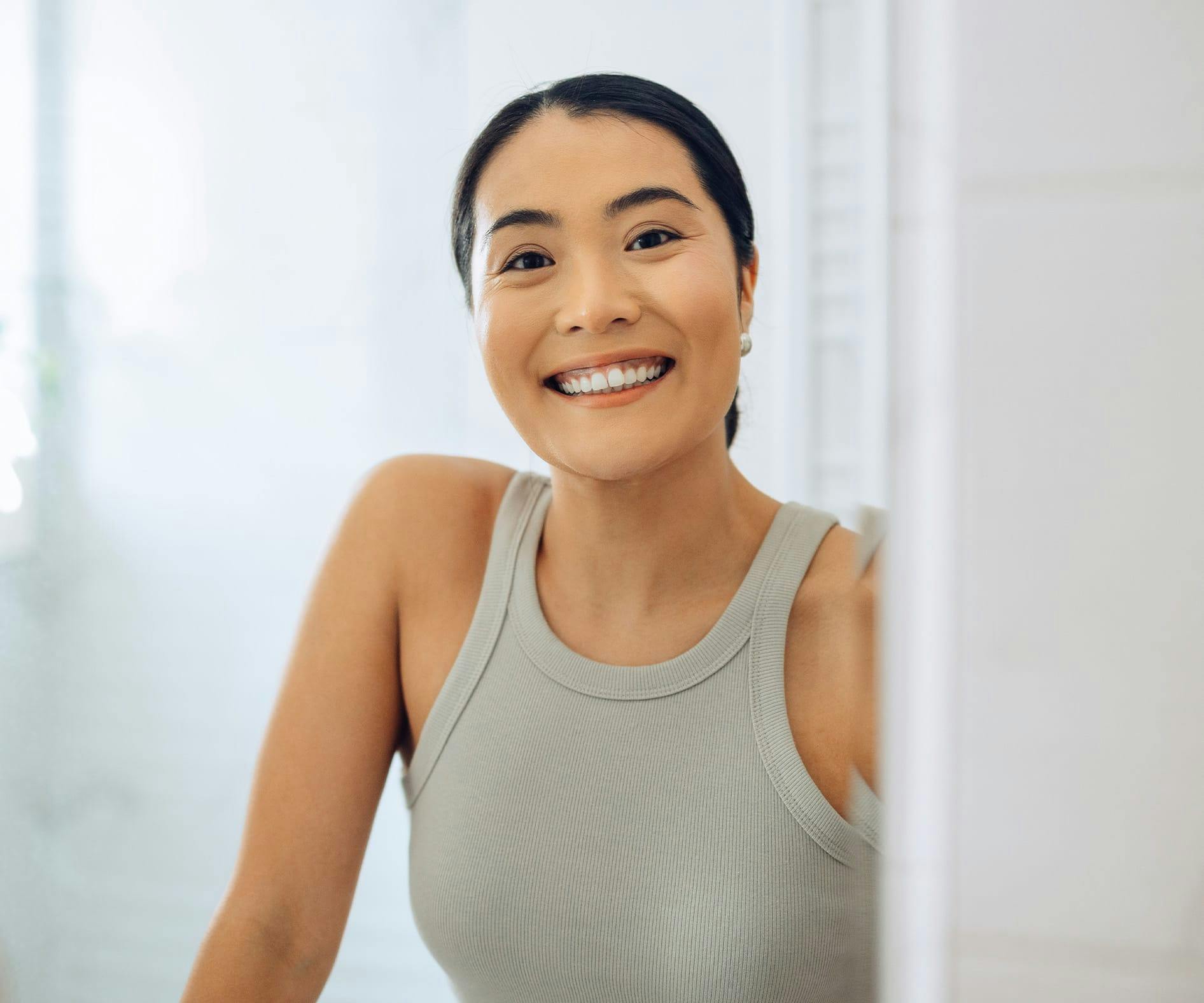 smiling woman in grey tank top looking at mirror in bathroom