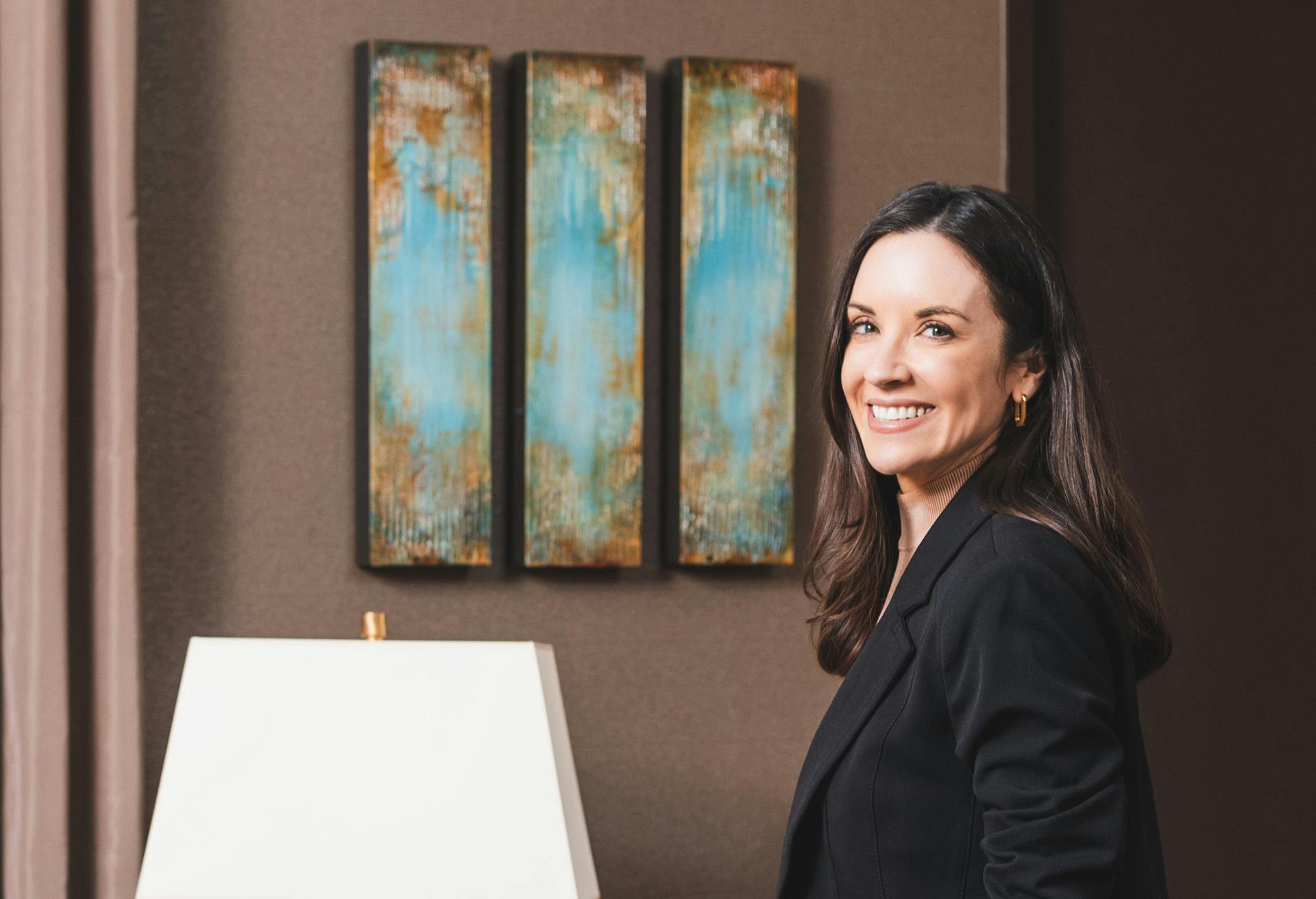 Woman in a black blazer smiling in front of a piece of wall art