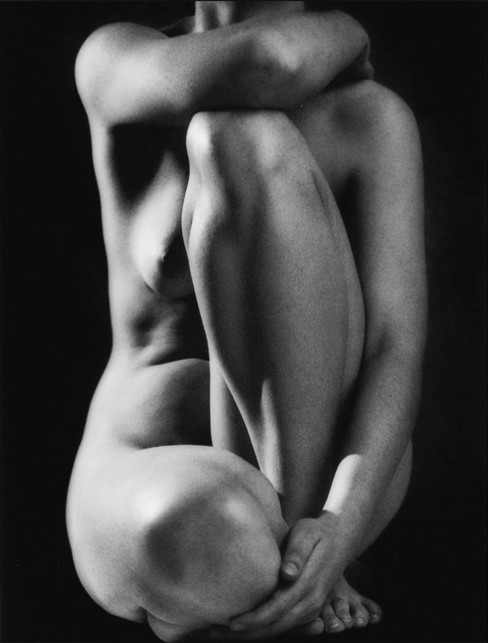 Nude woman sitting down, crossing her legs and arms over herself