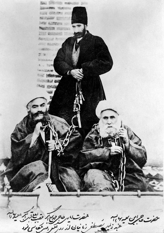 Hands of the Cause Hají Ákhúnd and Hájí Amín (seated), in chains and stocks with jailor standing above, Iran, 1890s