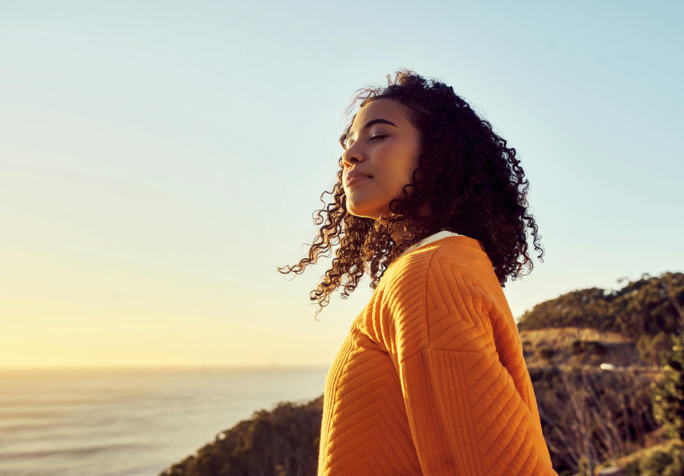 Woman Standing on a Hill by the Ocean with an Orange Sweater and Curly Dark Hair