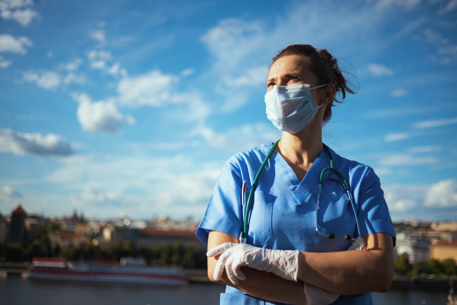 Health professional wearing scrubs, gloves, and a mask standing in front of a blue sky