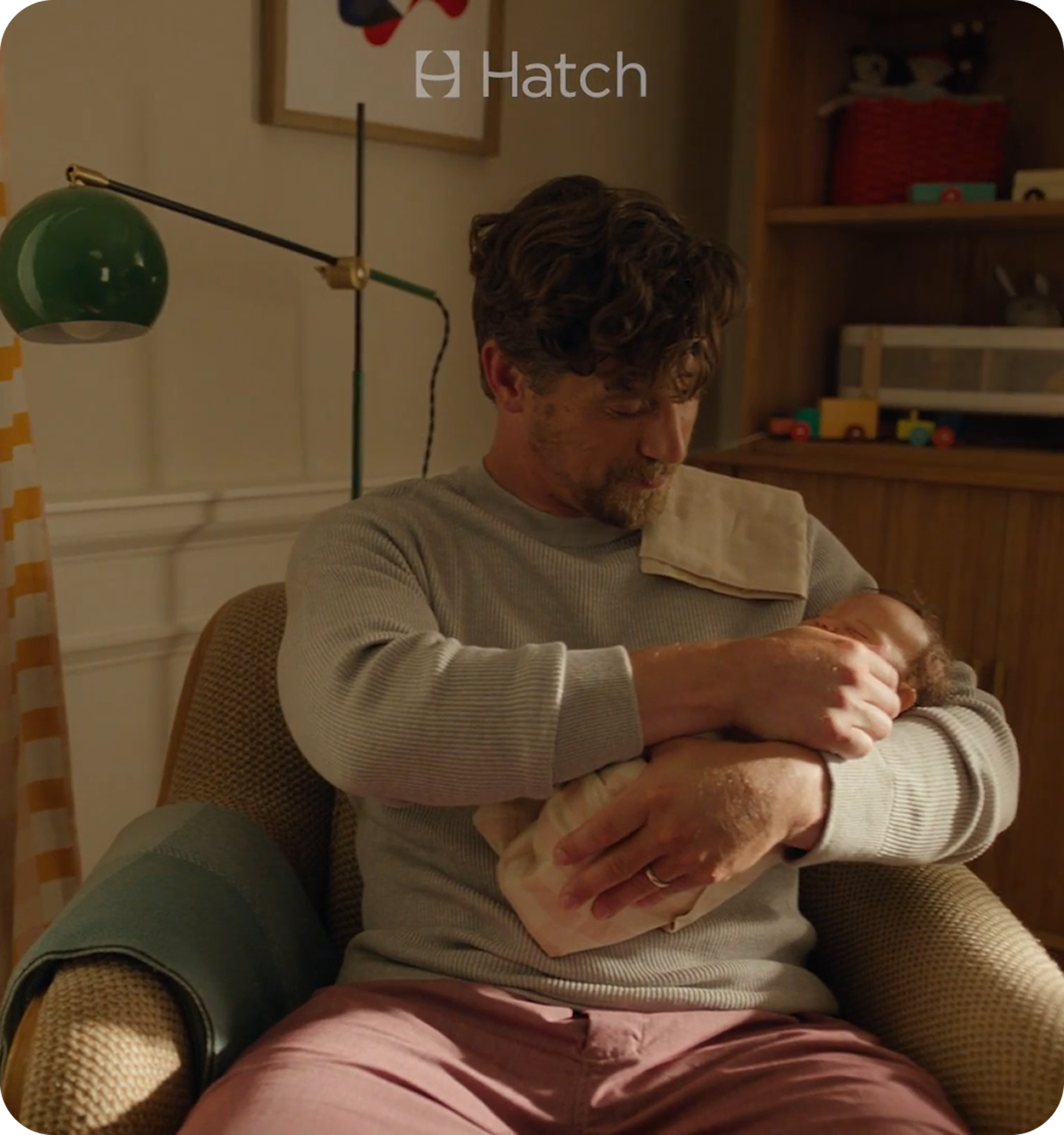 A parent cradling a swaddled newborn in a nursery armchair, lit by a green desk lamp