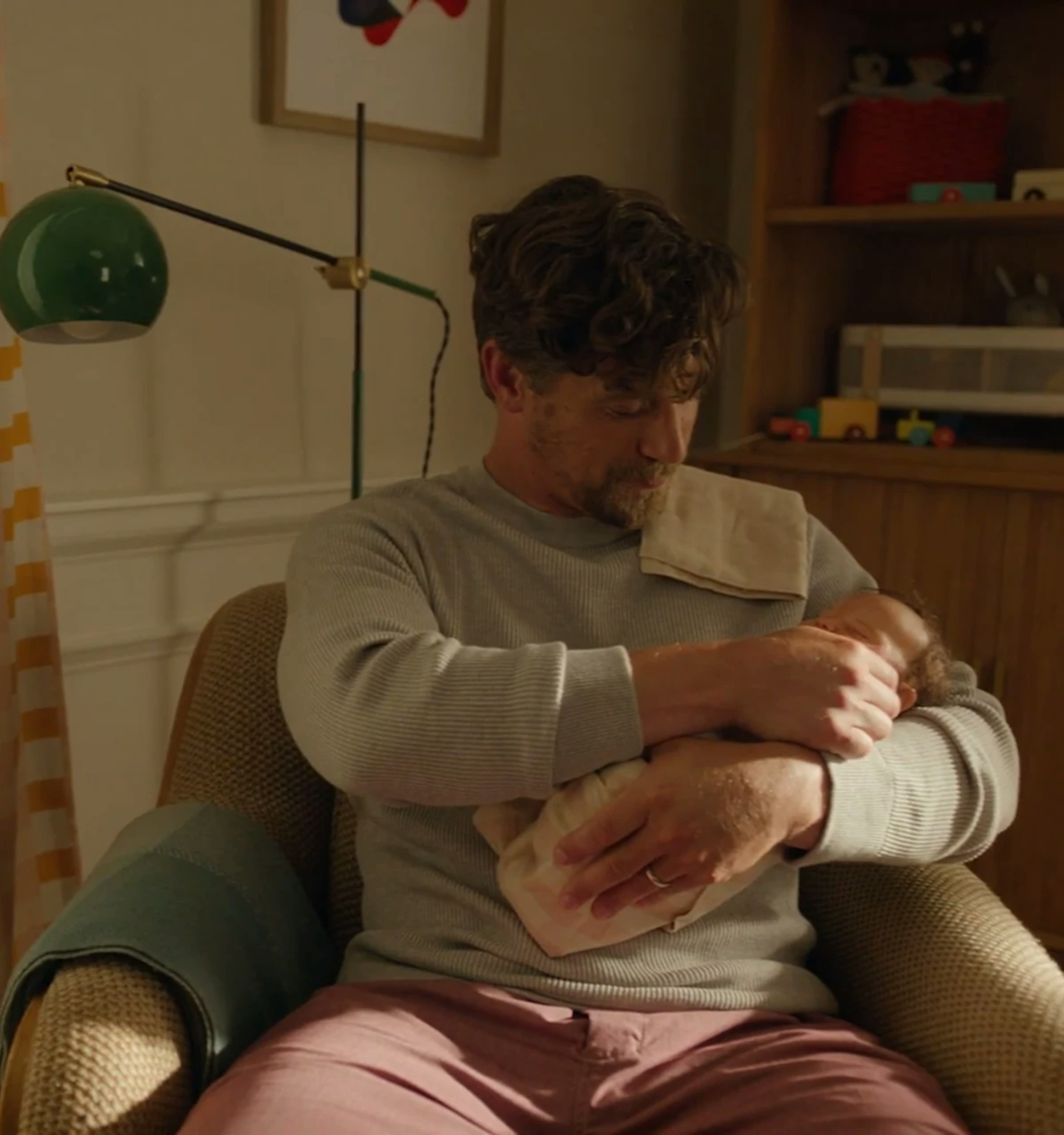 A parent cradling a swaddled newborn in a nursery armchair, lit by a green desk lamp