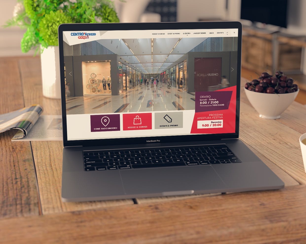 Un laptop è posizionato su un tavolo di legno, mostrando una pagina web di un centro commerciale con varie sezioni, mentre una tazza di tè e una ciotola di frutta appaiono accanto, creando un'atmosfera rilassata.