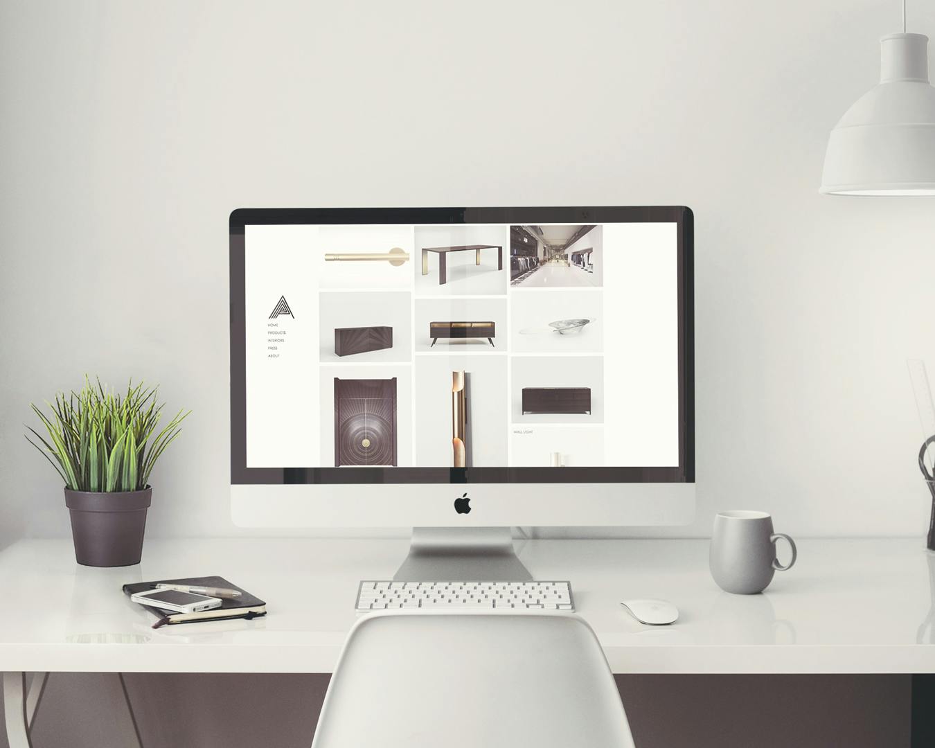 Una scrivania moderna e minimalista con un computer iMac con schermo acceso, una pianta in un vaso nero, una tazza bianca, una lampada sospesa e vari articoli di cancelleria, il tutto in un ambiente luminoso e ordinato.